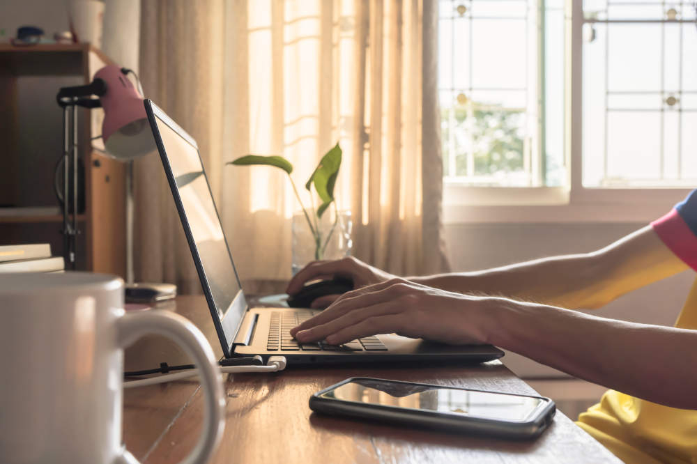 Un ordinateur posé sur un bureau avec une plante verte, un téléphone, une tasse. On voit les mains de la personne qui travaille.