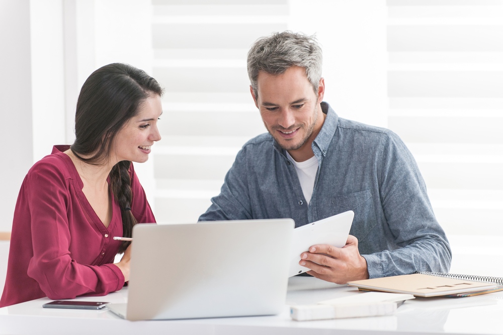 couple en train de lire quelque chose sur une tablette dans une pièce toute blanche avec un ordinateur posé sur la table devant eux